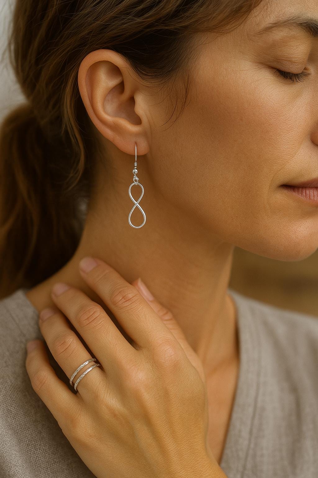 Woman wearing silver infinity earrings and ring, with a neutral background