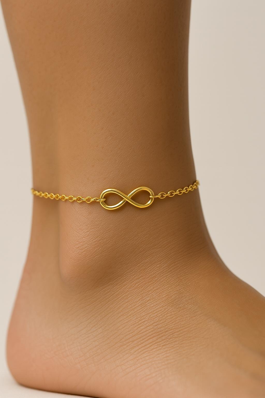 Gold infinity anklet on a person's ankle against a neutral background