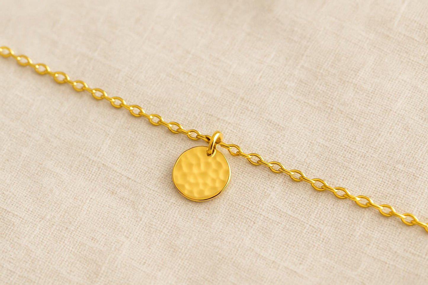 Gold bracelet with a round, textured charm on a beige fabric background