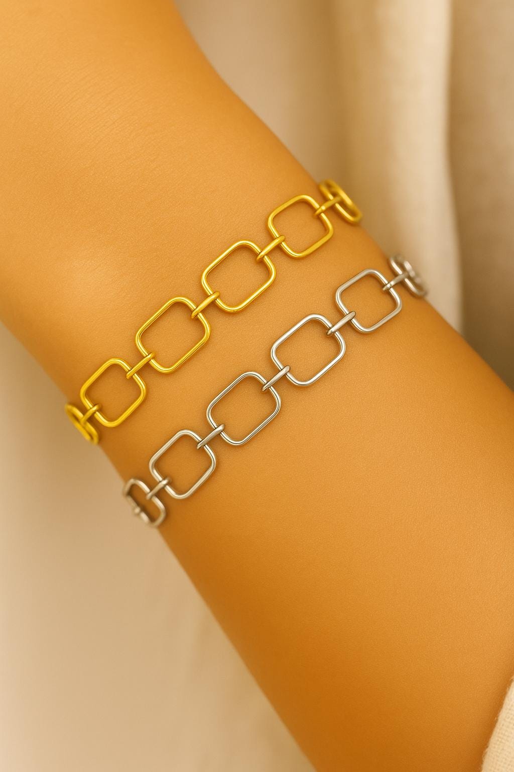 Two chain-link bracelets, one yellow and one silver, on a person's wrist against a beige background.