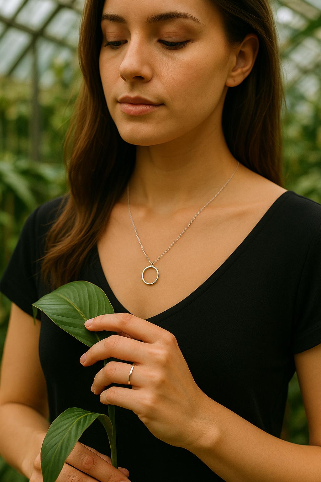 Woman wearing a necklace with a circular pendant, holding a leaf in a natural setting.