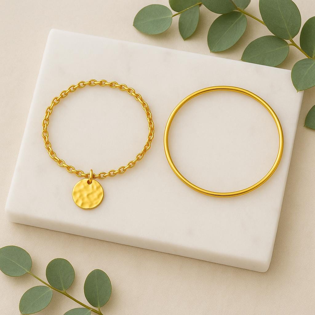 Two gold bracelets on a white marble surface with green leaves.