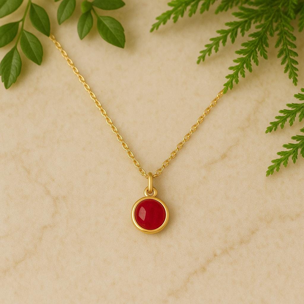 Gold necklace with a red pendant on a beige background with green leaves.