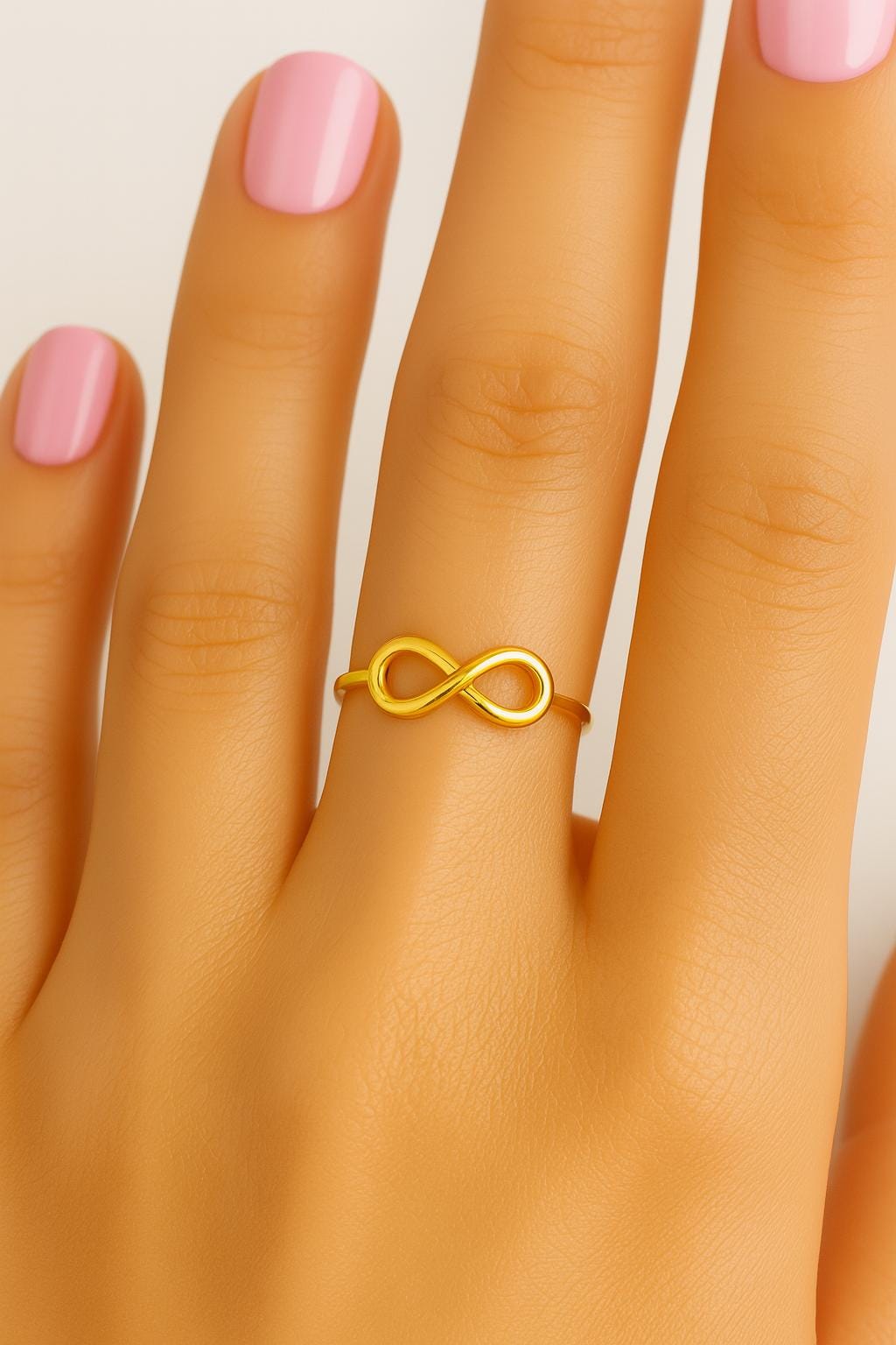 Gold infinity ring on a hand with pink nail polish against a neutral background