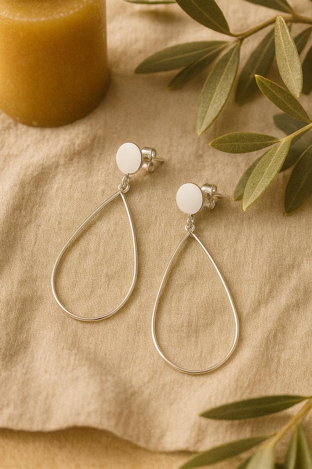 Teardrop-shaped silver earrings on a beige fabric background with green leaves.