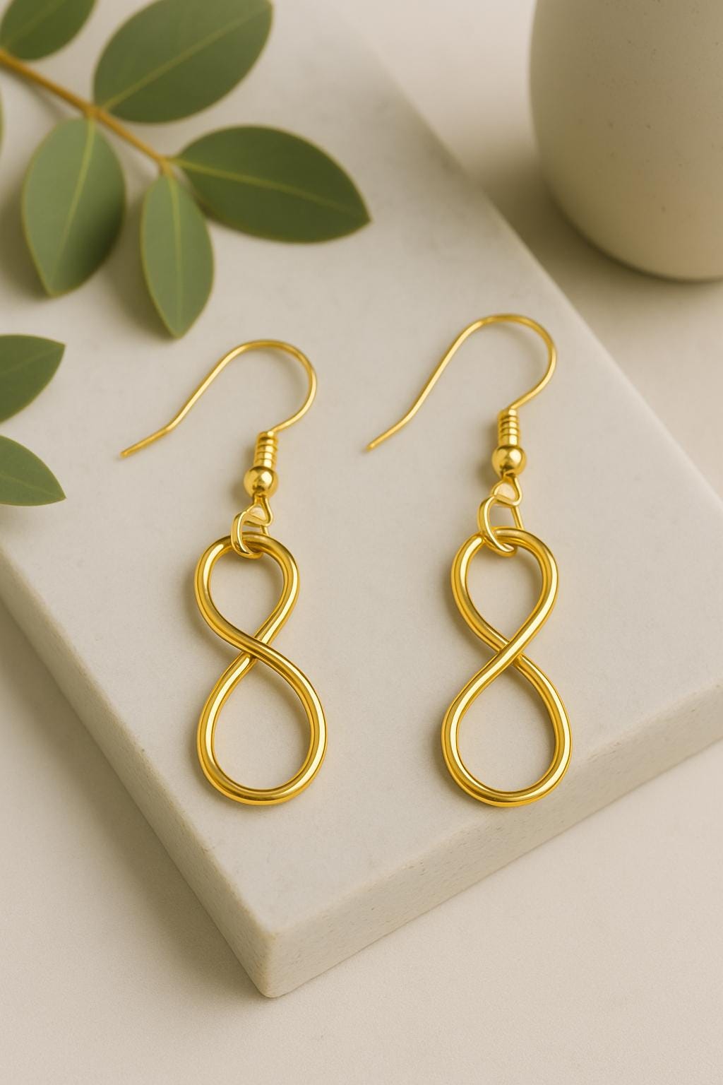Gold infinity-shaped earrings on a beige surface with green leaves.