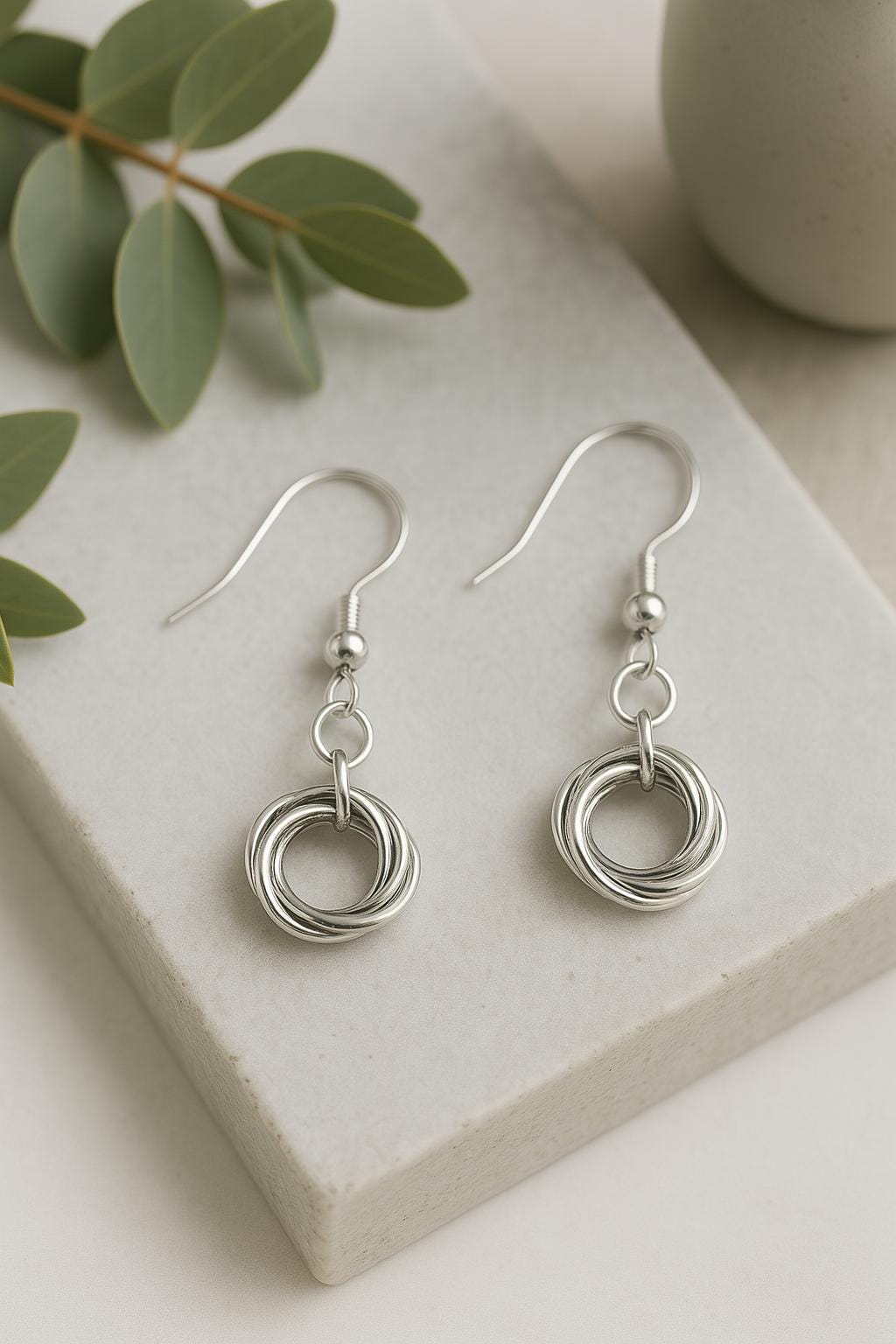 Silver earrings on a stone surface with green leaves and a white vase in the background
