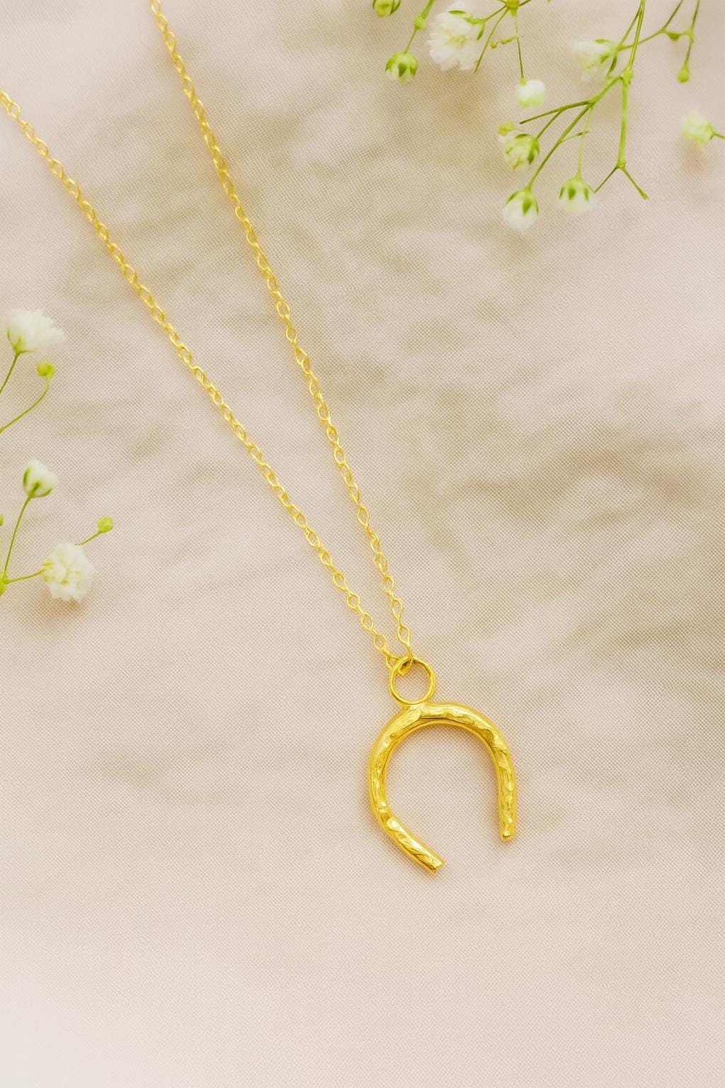 Gold hammered horseshoe necklace on a beige background with small white flowers