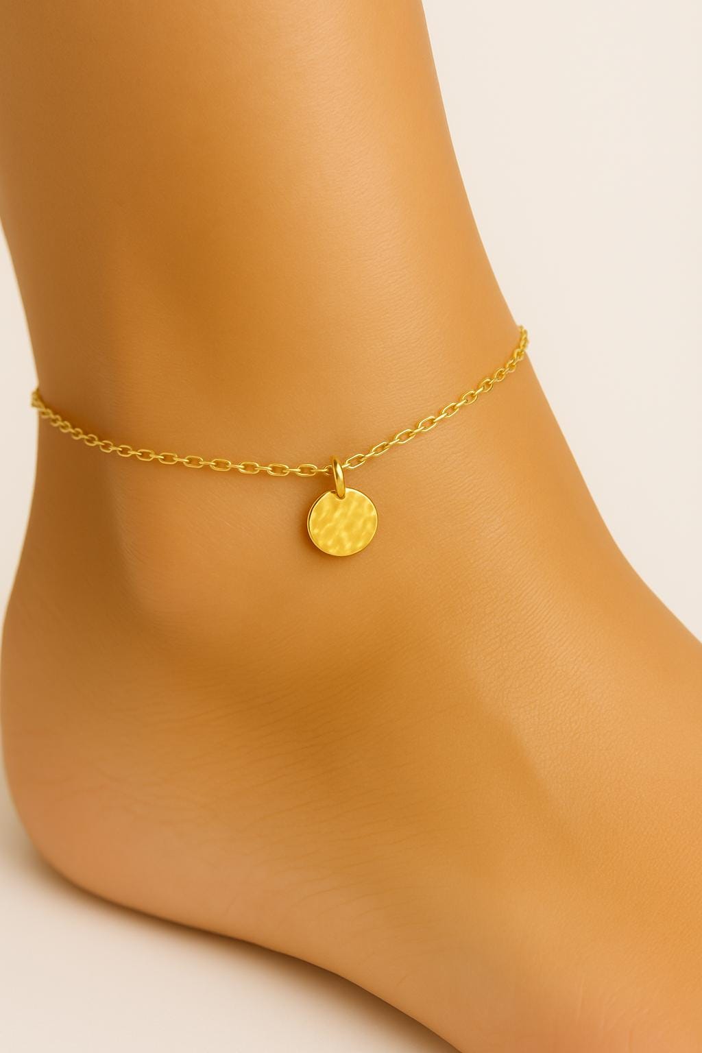 Gold anklet with a round charm on a foot against a neutral background