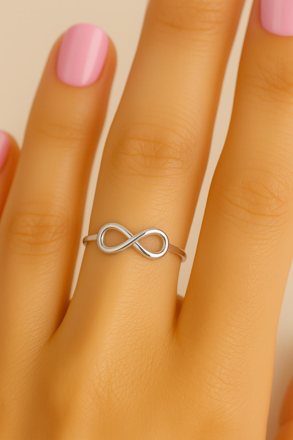 Silver infinity ring on a hand with pink nail polish against a beige background