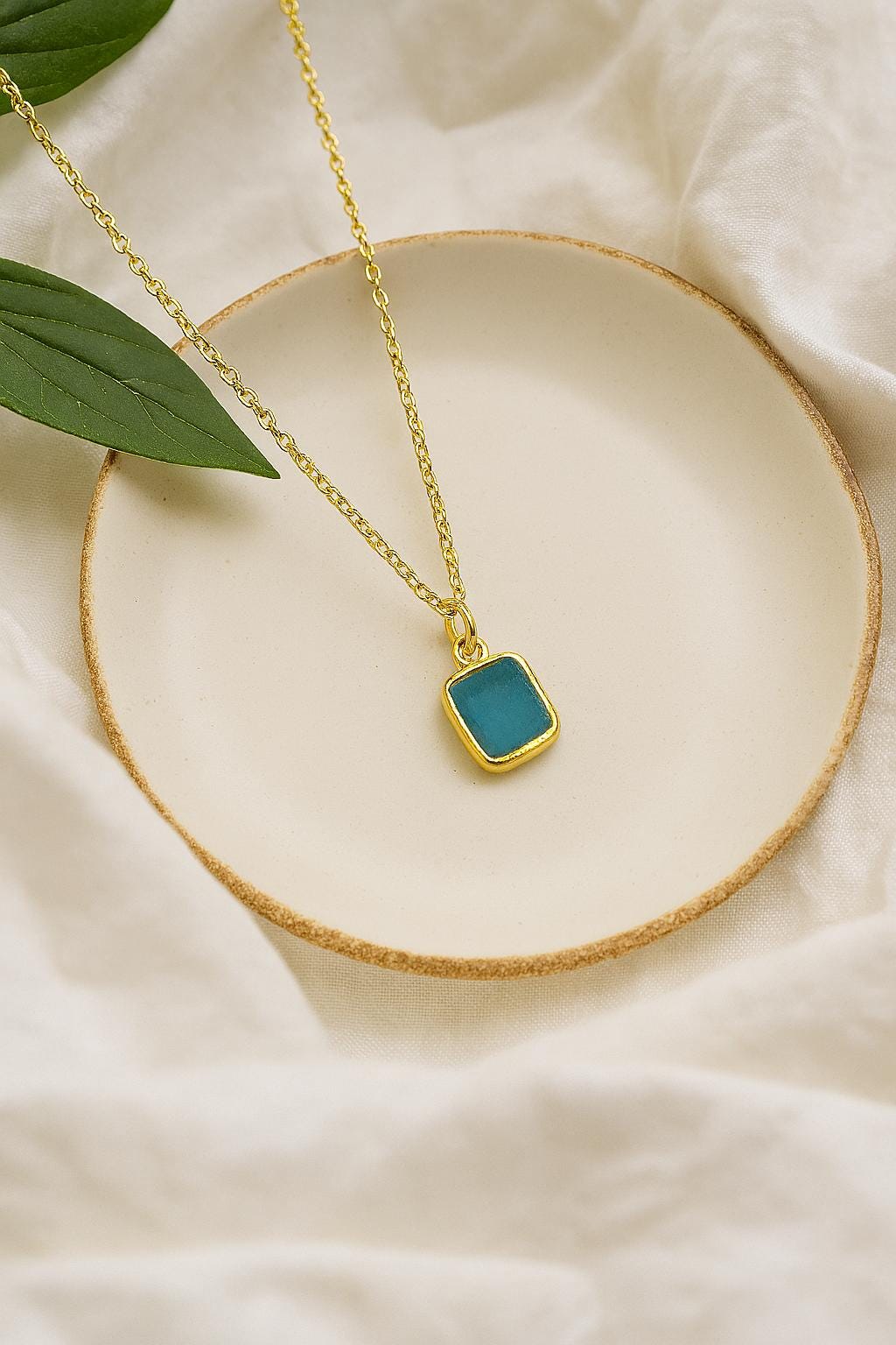 Gold necklace with a blue pendant on a beige surface with green leaves.