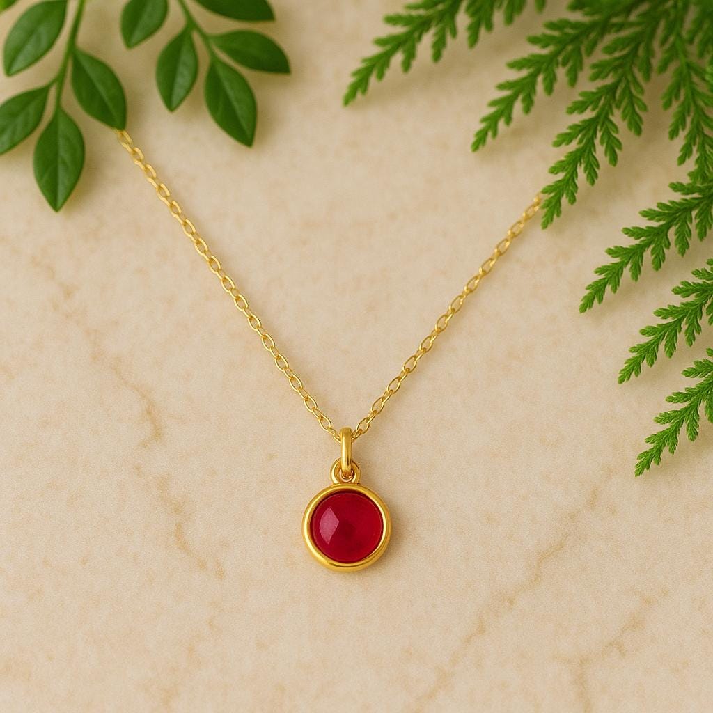 Gold necklace with a red pendant on a beige background with green leaves.