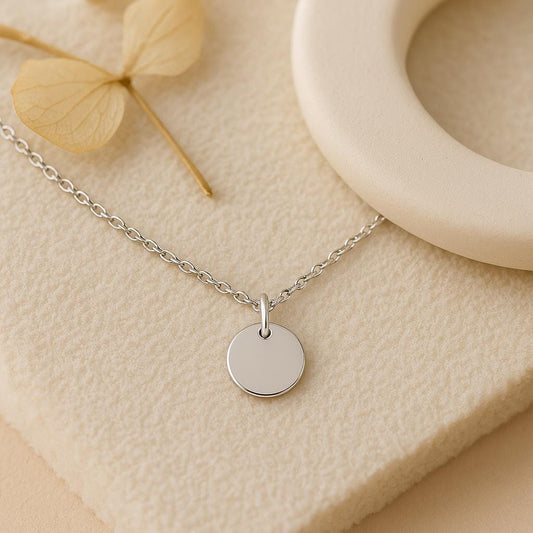 Silver necklace with a round pendant on a beige surface with dried leaves and a wooden ring.