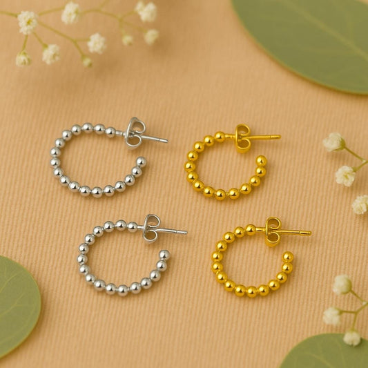 Two pairs of bead hoop earrings, one silver and one gold, on a beige background with green leaves and white flowers.
