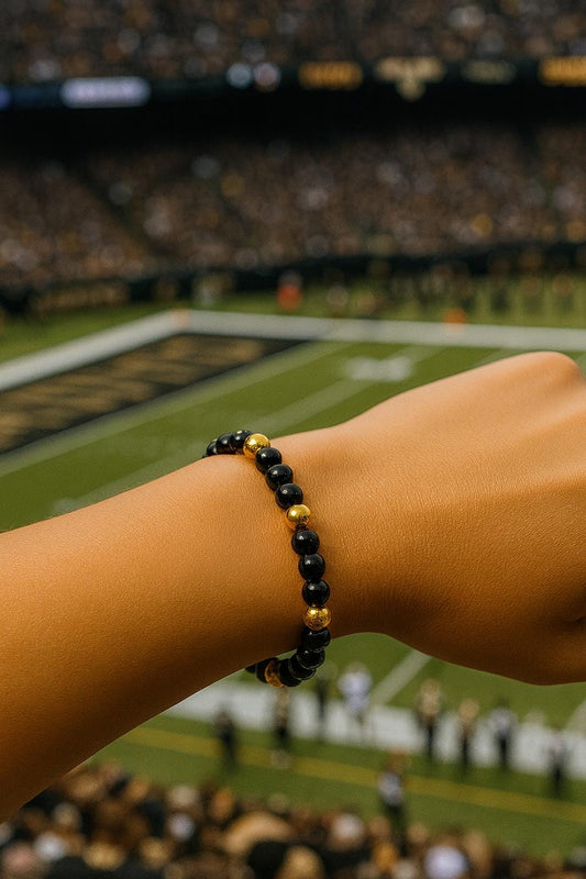 Person wearing a black and gold beaded bracelet at a sports event with a blurred stadium background