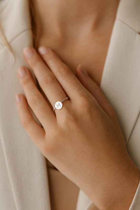 Hand wearing a ring with a visible 'M' on a neutral background