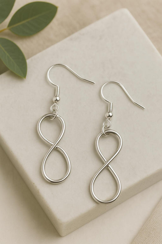 Silver infinity-shaped earrings on a beige surface with green leaves.