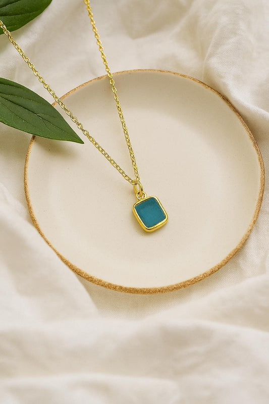 Gold necklace with a blue pendant on a beige surface with green leaves.